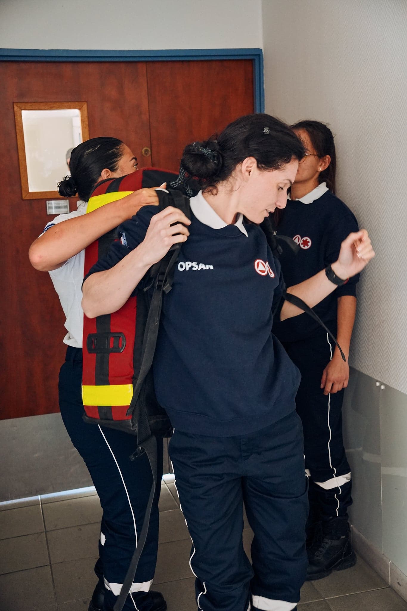 Bénévoles OPSAN s'équipant avant intervention, une équipière aide à passer le sac d'intervention rouge